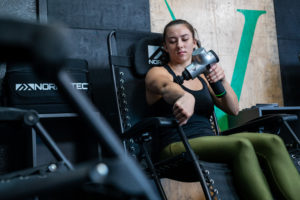 Invictus Athlete in the recovery area using the Hypervolt on her biceps.